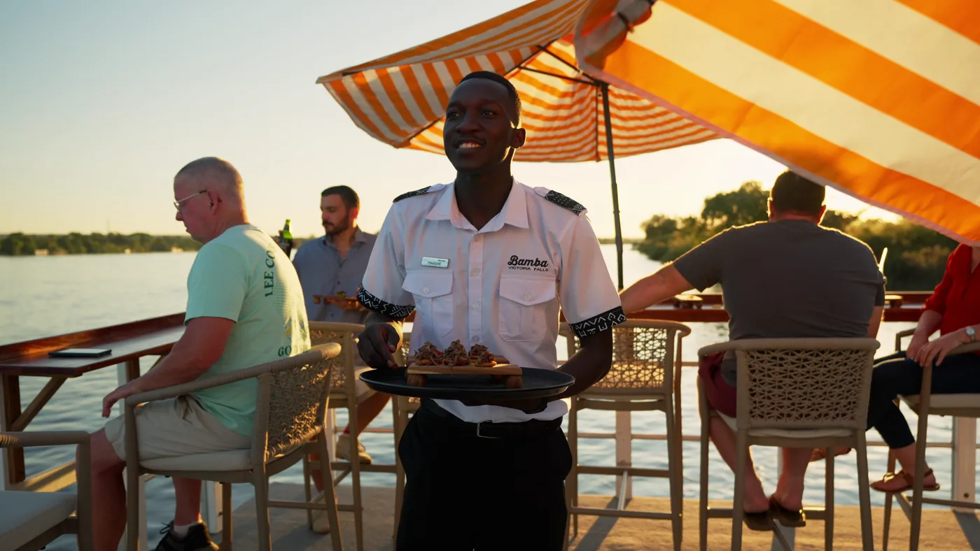 A Bamba crew member serving canapes on deck under striped umbrellas